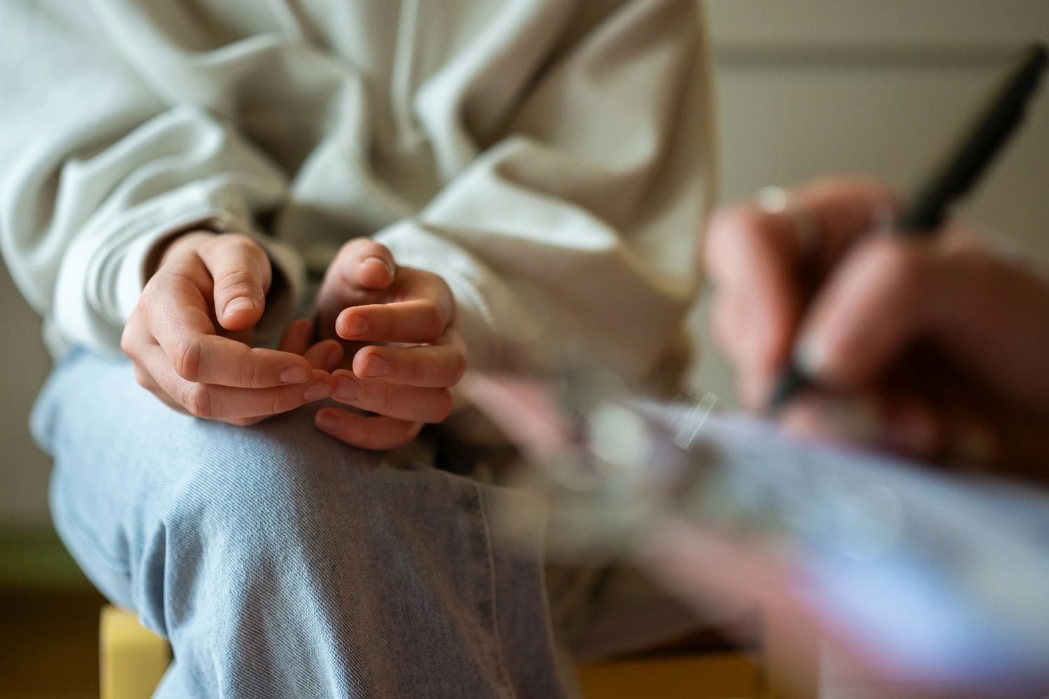 Person sitting with hands clasped, another writing on a clipboard, possibly in a counseling session.