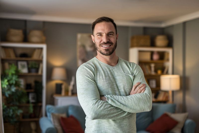 Portrait of a smiling mid-adult man with arms crossed, radiating confidence and contentment in a modern, stylish living room filled with cozy decor and natural light.
