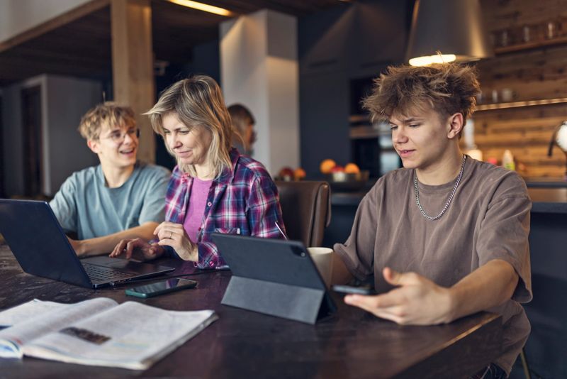 A mother and three teenagers sitting around a table in a large open kitchen. The mother is focused on her laptop, working, while the teenagers are engaged in a study session. The mother is wearing a plaid shirt, while the teenagers are dressed in casual clothing. The group is focused on their activities, with the mother working on her laptop and the teenagers using a tablet, a laptop, and few open books - collaborative and productive environment among the family members.Shot with Canon R5