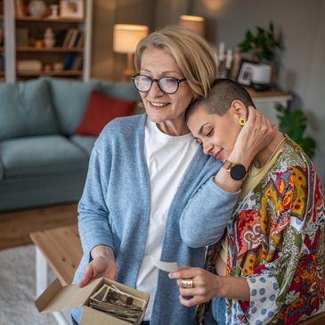Two women share a tender moment looking at old photographs in a cozy living room.