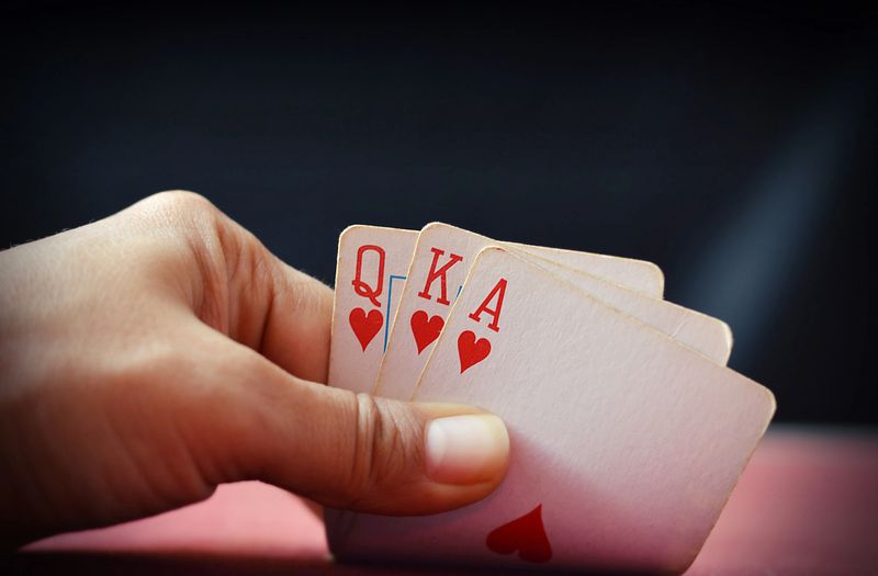 A hand picking up, giving or secretly showing three playing cards, ace, queen and king of heart over red poker table against black background for Royal Flush, in Poker, Gambling, casino related backdrops with copy space.