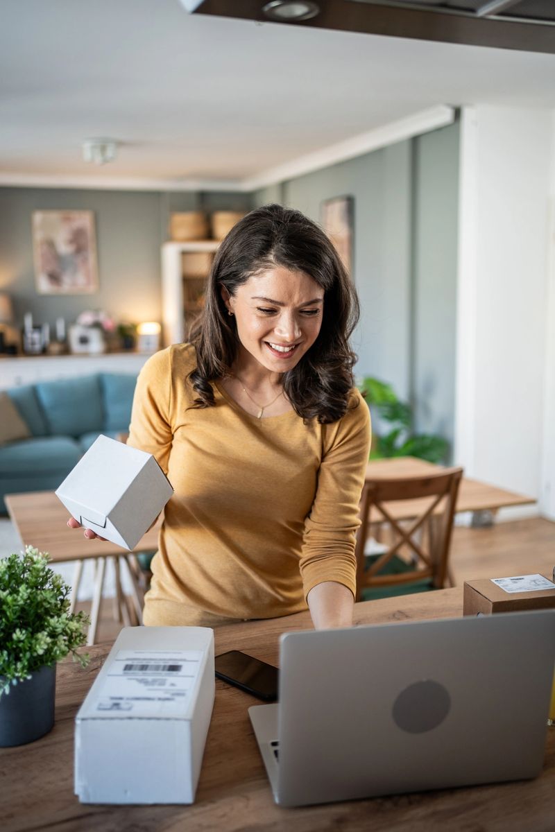 Young woman smiling while presenting a white box in front of her laptop during a lively livestream at home, engaging with her followers in a cheerful and enthusiastic manner.