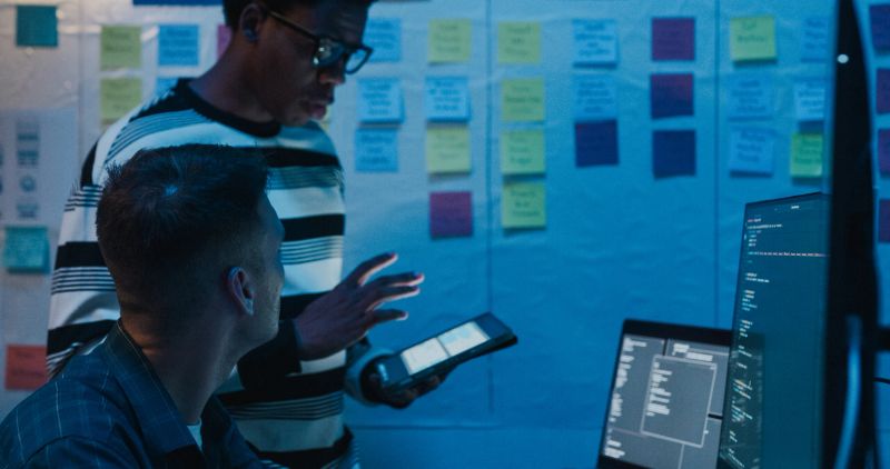 Group of IT professionals collaborating on network security and data analysis using digital tablet and laptop in office at night. Software developer, artificial intelligence and programming concept.