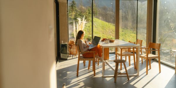 Woman working on a laptop in a sunlit room with large windows and wooden furniture.