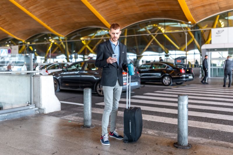 Young businessman using his smartphone while waiting for a taxi at the airport, holding a reusable water bottle