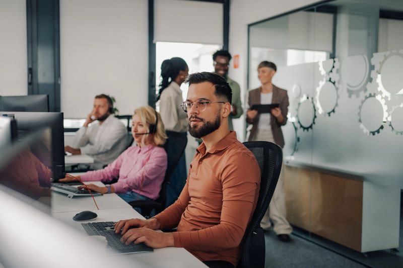 In a bustling call center, a male operator wearing glasses concentrates on his computer, diligently typing while colleagues assist customers and managers discuss strategies