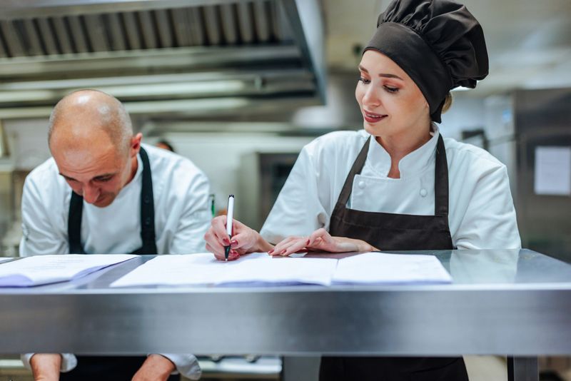 Two chefs collaborating in a bustling restaurant kitchen, diligently taking notes on documents while planning and organizing their culinary tasks