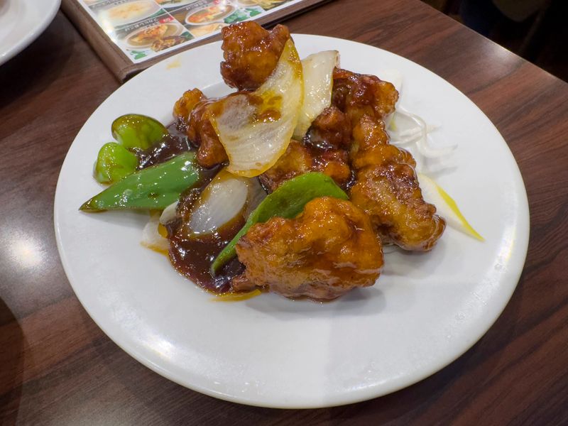 A top-down shot of a white plate filled with sweet and sour pork (咕咾肉/酢豚) at an all-you-can-eat restaurant in Yokohama Chinatown. The dish features crispy fried pork pieces coated in a glossy, reddish-brown sweet and sour sauce, accompanied by bell peppers and onions. The plate is placed on a wooden table, suggesting a casual dining setting. The image captures a classic Chinese dish served in a Japanese context.