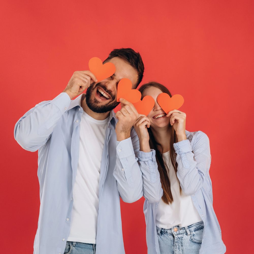 Happy couple playfully holding paper hearts over their eyes against a red background.