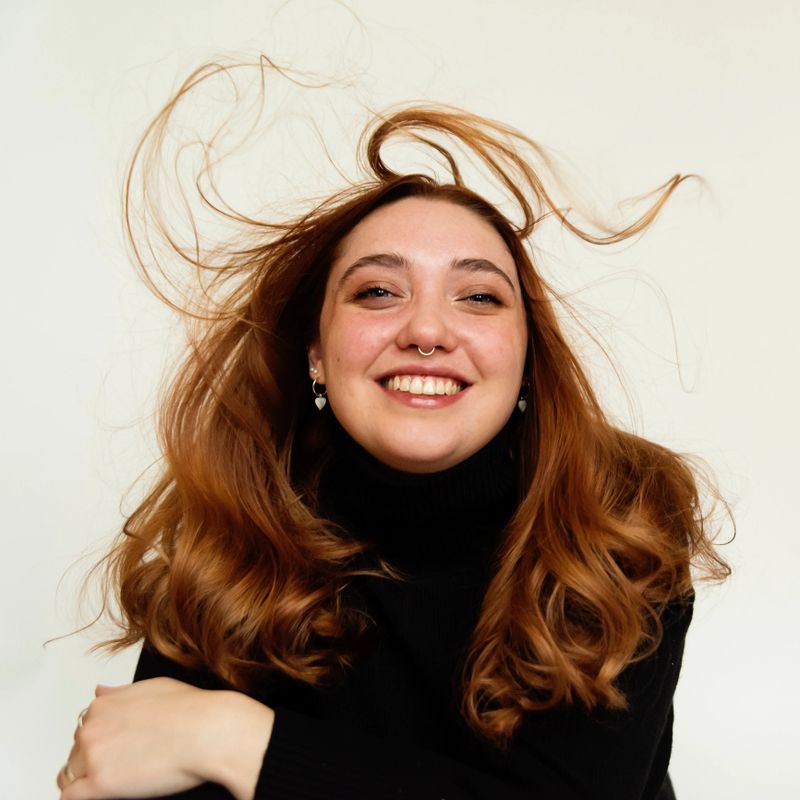 Portrait of redhead woman in her twenties on white background. She is wearing a black turtleneck pullover, and is looking at the camera with her very blue eyes. Wind in her hair. Square studio headshot shoot in natural light. This was taken in Montreal, Quebec, Canada.