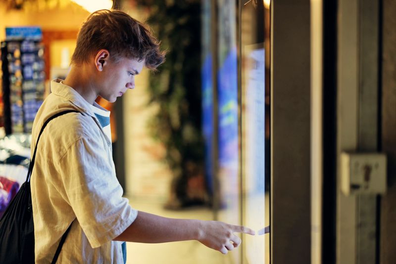 A teenage boy interacting with a vending machine in an underground setting. The boy is wearing a casual outfit with a backpack slung over one shoulder. He is intently selecting a drink from the vending machine's touchscreen interface. The environment around him includes tiled walls and somewhat dim lighting typical of underground spaces.
Shot with Canon R5