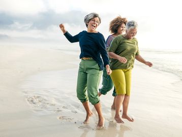 Three joyful women jumping and laughing on a sandy beach near the ocean.