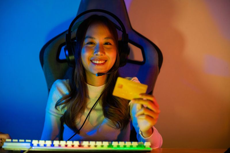 Asian gamer playing online video game and holding a credit card for payment game online, young woman in gaming headphones using computer for playing games at home, broadcast streaming live, Front view