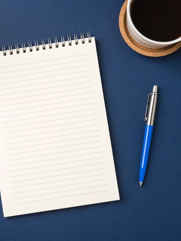 Empty notebook, blue pen, and coffee cup on blue surface.