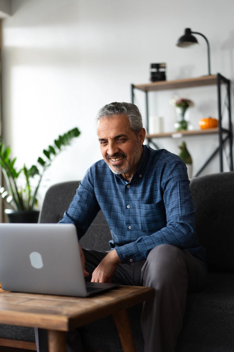 senior adult man smiling while working on a laptop in a comfortable living room setting with modern decor