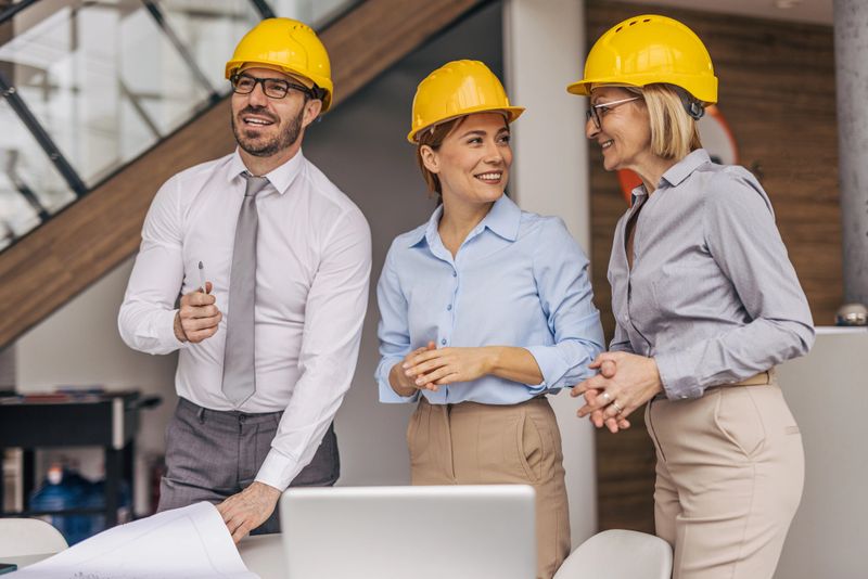 Architects and engineers wearing hard hats are standing around a table, discussing blueprints and smiling while working together on a project