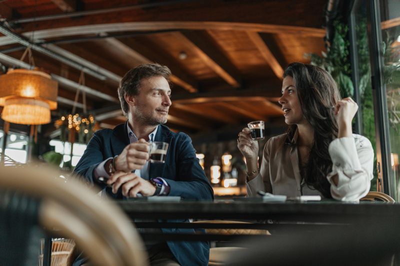 A stylish couple enjoys an intimate conversation over coffee at a modern cafe, surrounded by warm lighting and lush greenery, blending comfort with sophistication.