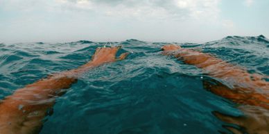 Hands reaching out of deep blue ocean water under a cloudy sky.