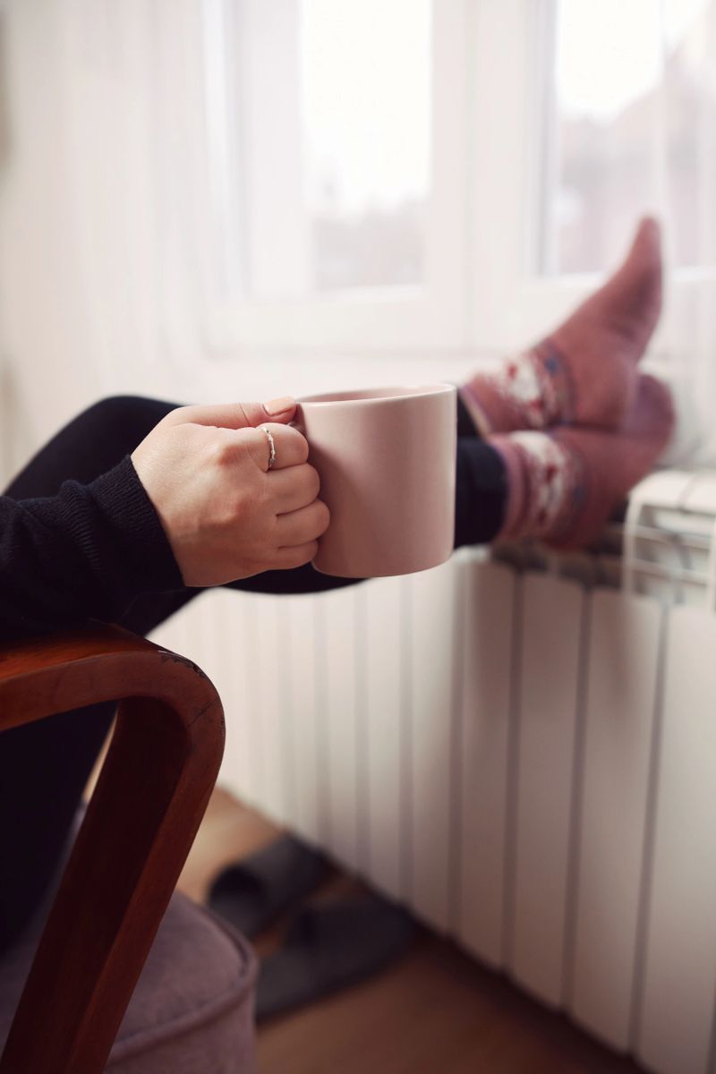 Woman drinking hot tea with her feet with socks on a home heating radiator on a cold winter day outdoors.