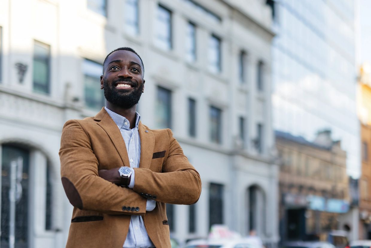 Confident man in a brown blazer smiling outdoors in an urban setting.