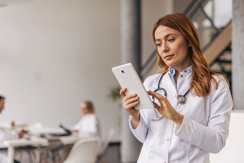 Confident female doctor wearing a lab coat and stethoscope, engaging with a digital tablet in a modern hospital, while colleagues collaborate and work in the background