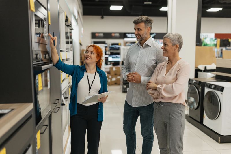 A saleswoman guides two customers as they explore modern kitchen appliances, showcasing various features and helping with their purchasing decisions in a retail setting.