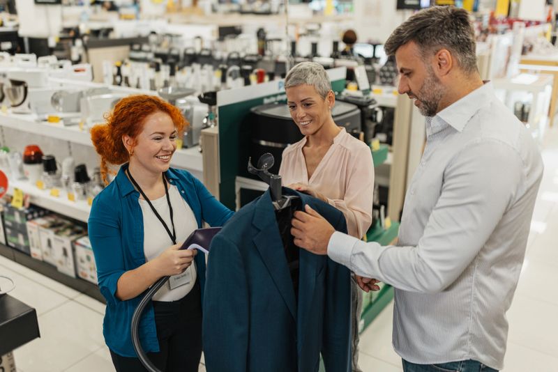 A knowledgeable salesperson displays a suit while explaining features to customers, showcasing excellent customer service in a modern retail environment filled with products.