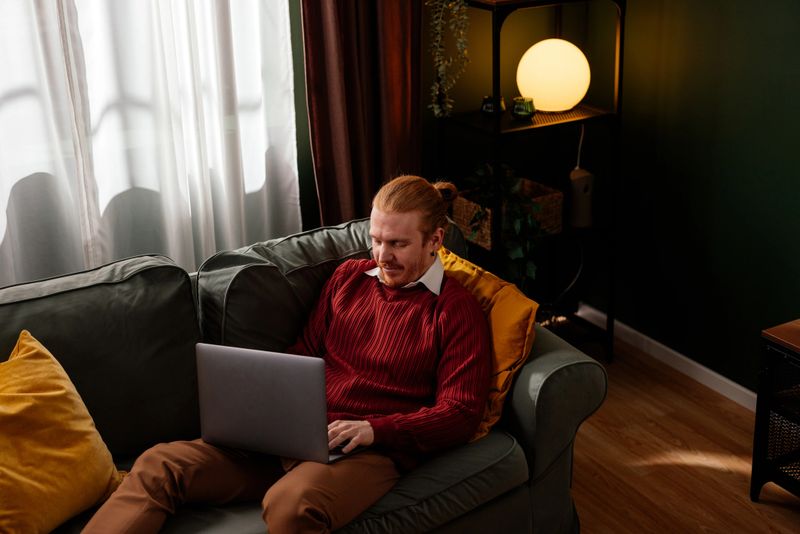 Redhead man using a laptop while sitting on a comfortable sofa, working from home in a cozy living room