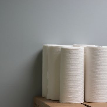 Stack of white paper towel rolls on a cardboard box against a gray wall.