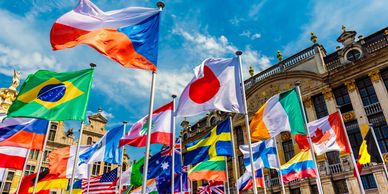 Multiple international flags waving in front of a historic European building under a blue sky.