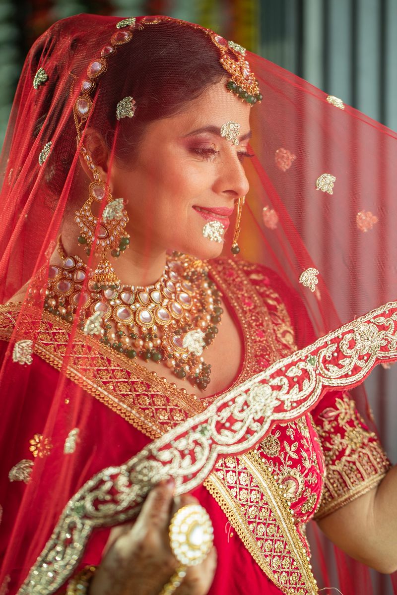 Portrait of beautiful bride dressed in traditional clothing and jewelries smiling and looking through red dupatta during wedding ceremony