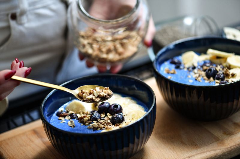 Female Arranging Blue Smoothie Bowls Adding Crunchy Granola on Top With Golden Spoon