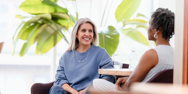 Two women engaged in a friendly conversation indoors with large green plants in the background.