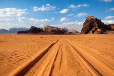 Tire tracks lead through a vast desert landscape with rocky formations under a blue sky.