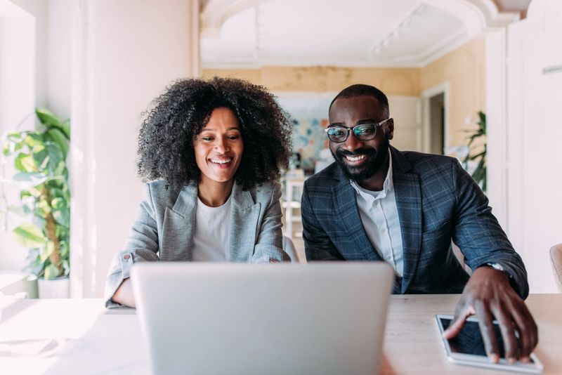 Shot of a two confident business persons sitting on a desk in the office and sharing ideas. Businessman and businesswoman in meeting using laptop and digital tablet and discussing business strategy. Business coworkers working together in the office.