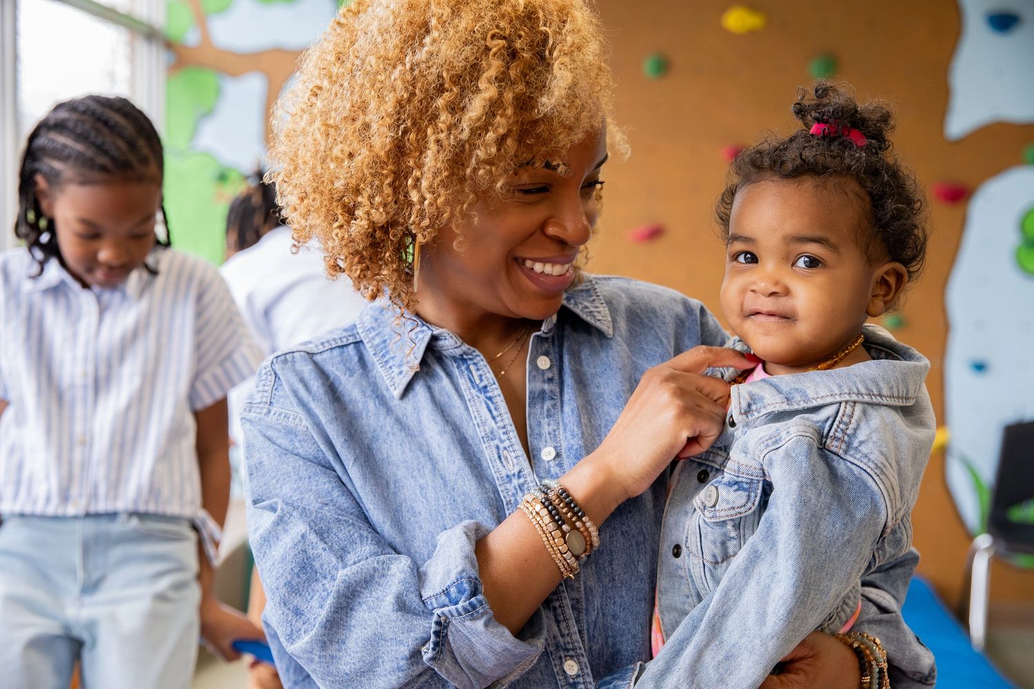A smiling woman holds a toddler wearing matching denim jackets indoors.