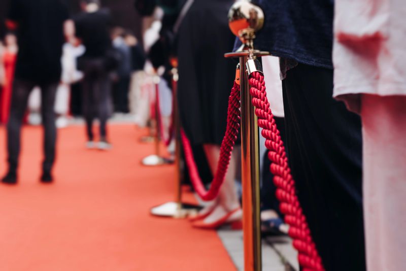 Red carpet with ropes and golden barriers on a luxury party entrance, cinema premiere film festival event award gala ceremony, wealthy rich guests arriving, outdoor decoration elements, summer day