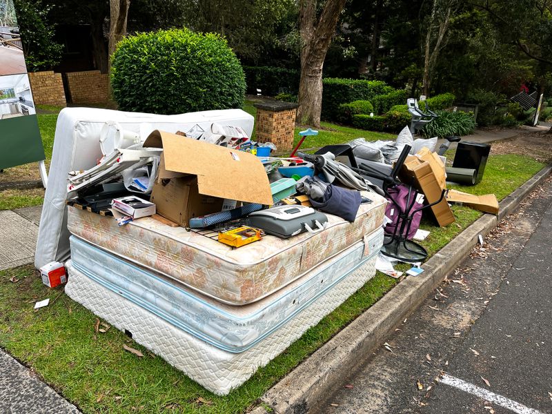 A large pile of rubbish set out on the curb for council collection in a suburban neighbourhood. The heap includes oversized items like flattened cardboard boxes, a worn couch with pillows, several mattresses stacked atop one another