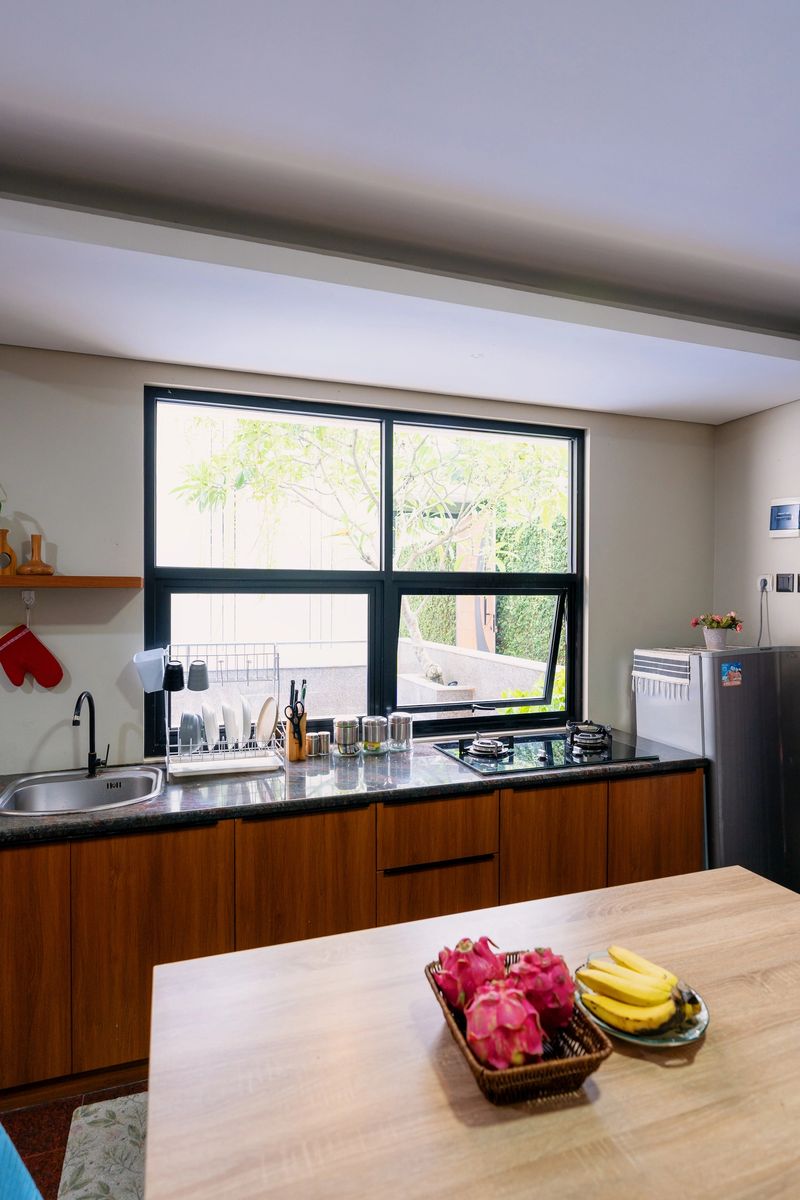 A warm and inviting open-concept kitchen and dining space with modern and minimalist decor eatures a sleek black countertop, a stainless steel sink, a wooden dining table with blue upholstered chairs, and a cozy living area with vibrant floral cushions.