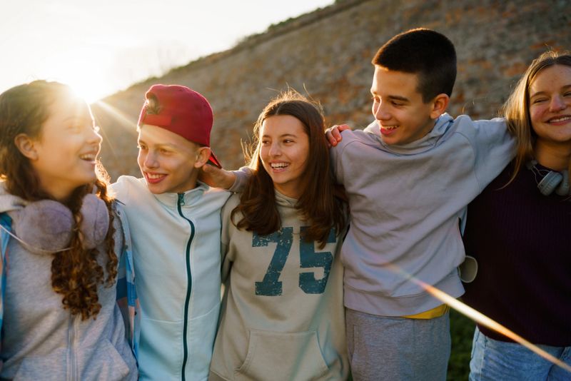 Happy teenage children are standing with their arms around each other outside.
