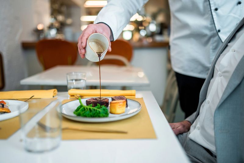 A refined fine dining setting featuring a chef pouring sauce over a gourmet dish, served with vibrant greens and a delicate potato side on a white plate.