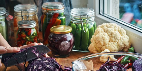 Person slicing purple cabbage with jars of pickled vegetables nearby.