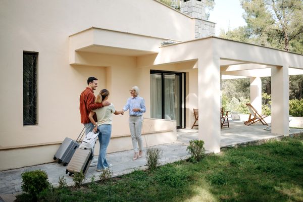 Couple arriving at a house, greeted by a woman with luggage.