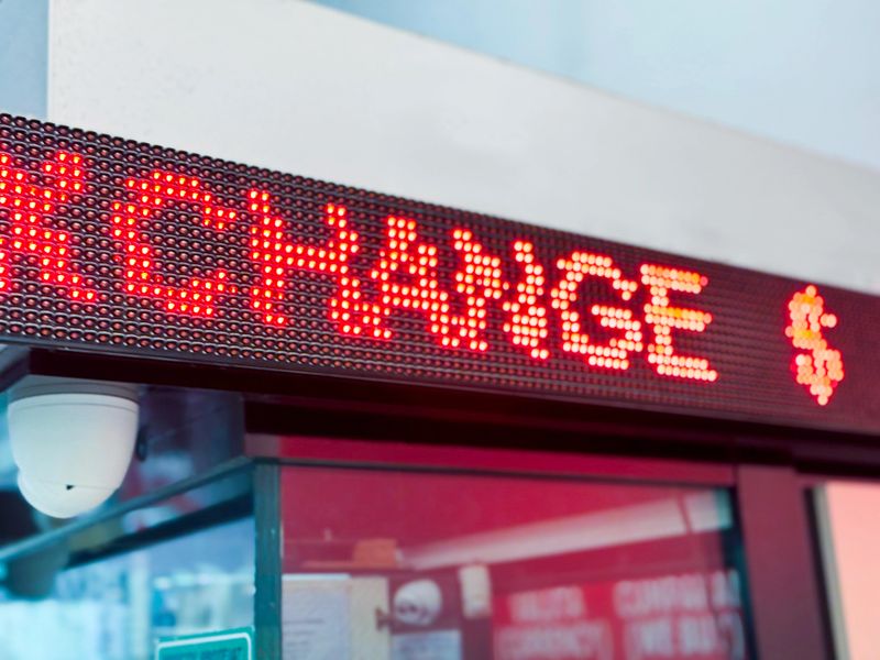 A close-up of a bright red LED display showing the word "EXCHANGE" with a dollar sign at a currency exchange booth, with a security camera mounted below and a glass window reflecting the surroundings.
