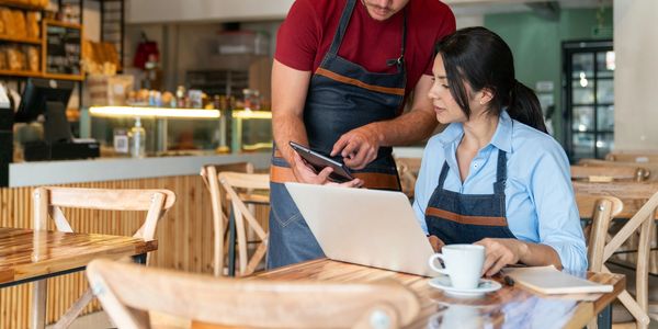 Two cafe workers discussing orders using a tablet and laptop.
