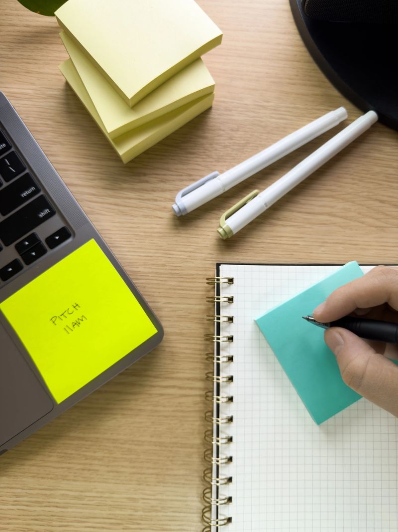 Top view of a home office desk setup with a stack of yellow Post-it® Dispenser Pop-up Notes 3301-5YW, pens, laptop with Super Sticky Post-it® reminder, and hand writing on notebook with Super Sticky Post-it® pad.
