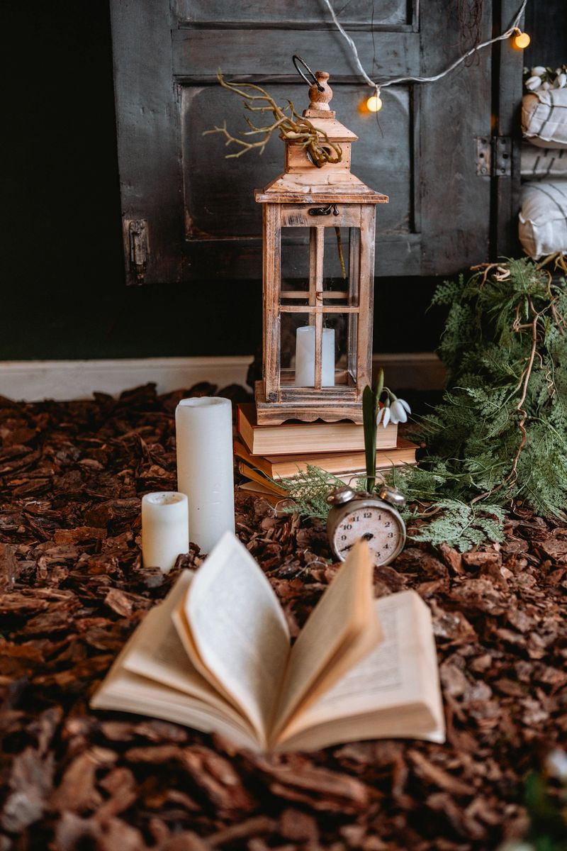 A rustic nook features an open book, lantern, candles, and greenery scattered around.