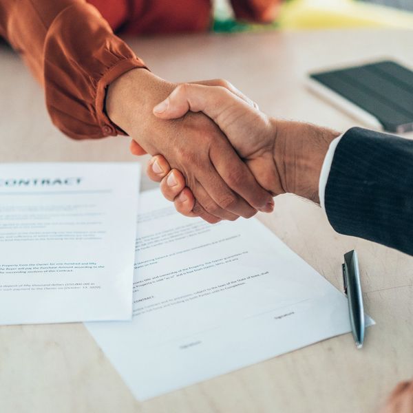 Two people shaking hands over signed contract documents on a table.