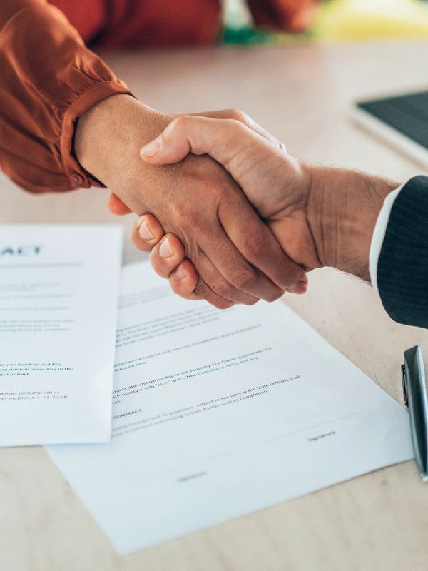 Two people shaking hands over signed contract documents on a table.