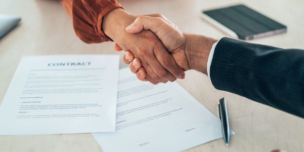 Two people shaking hands over signed contract documents on a table.